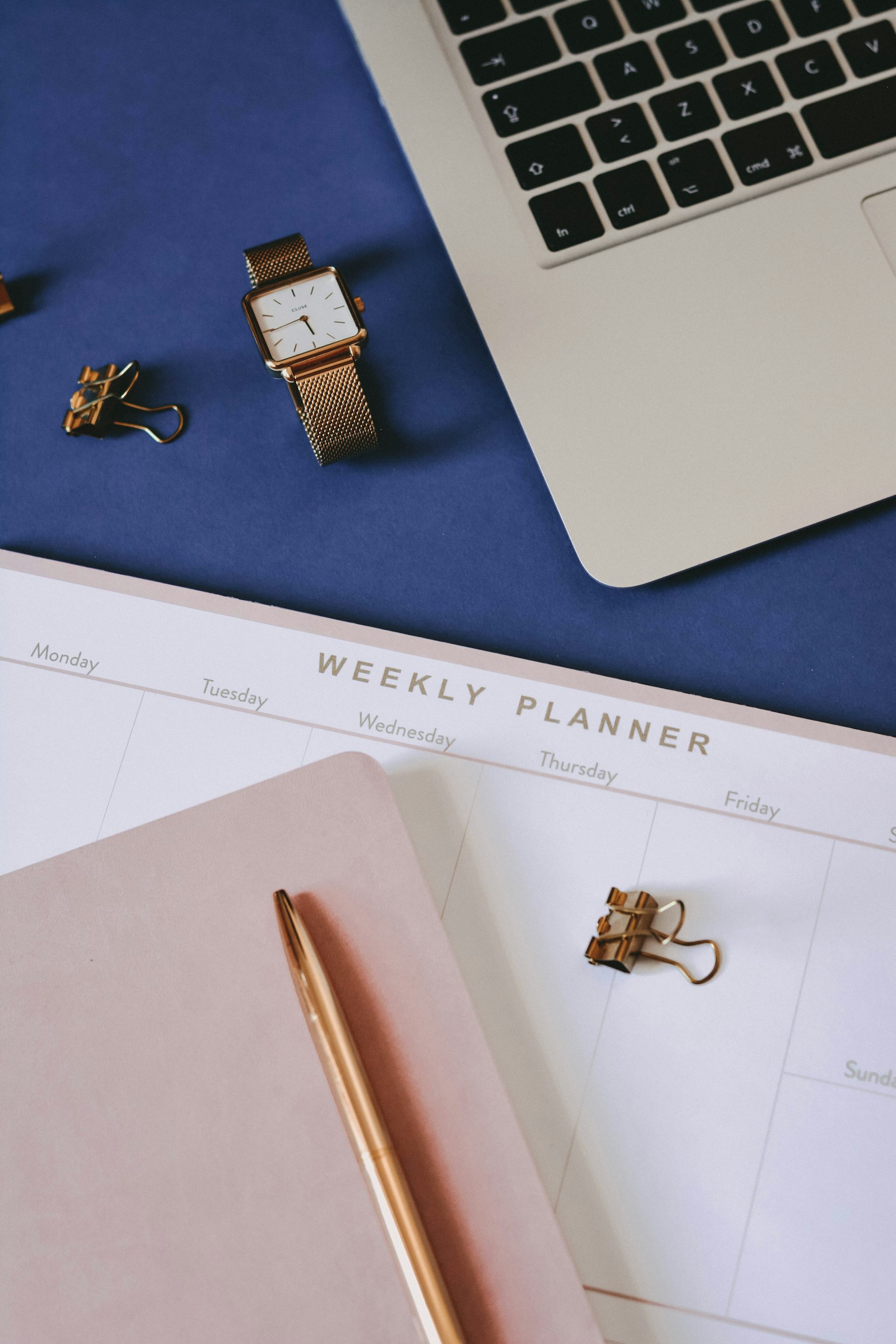 A weekly planner on a desk beside a laptop, watch and pen